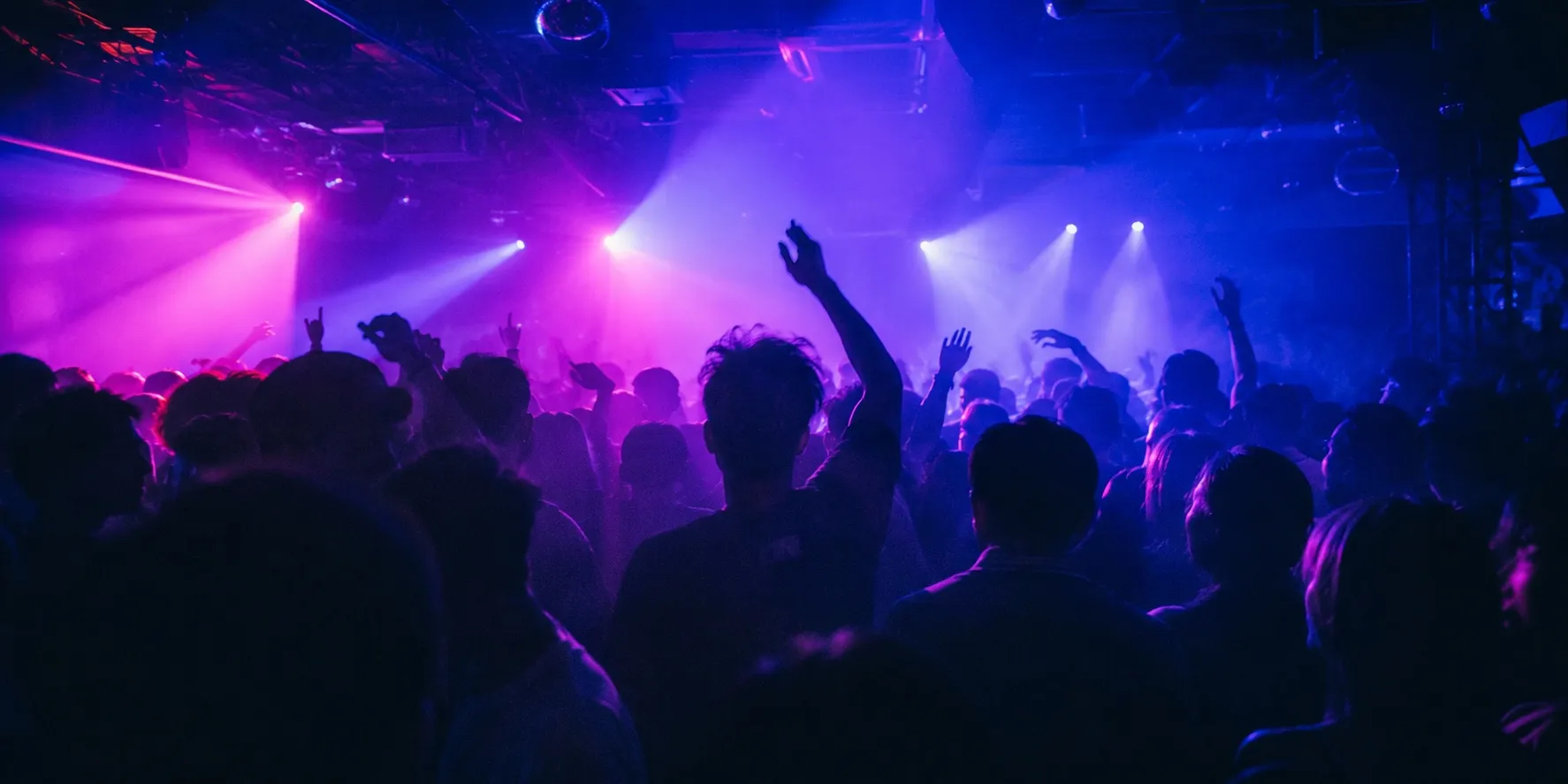 The dance floor of a nightclub at night, seen from behind the crowd. Dozens of dark silhouettes of heads and raised arms stand against beams of magenta, purple and blue light cutting through the haze from the ceiling. Nobody is holding a phone.