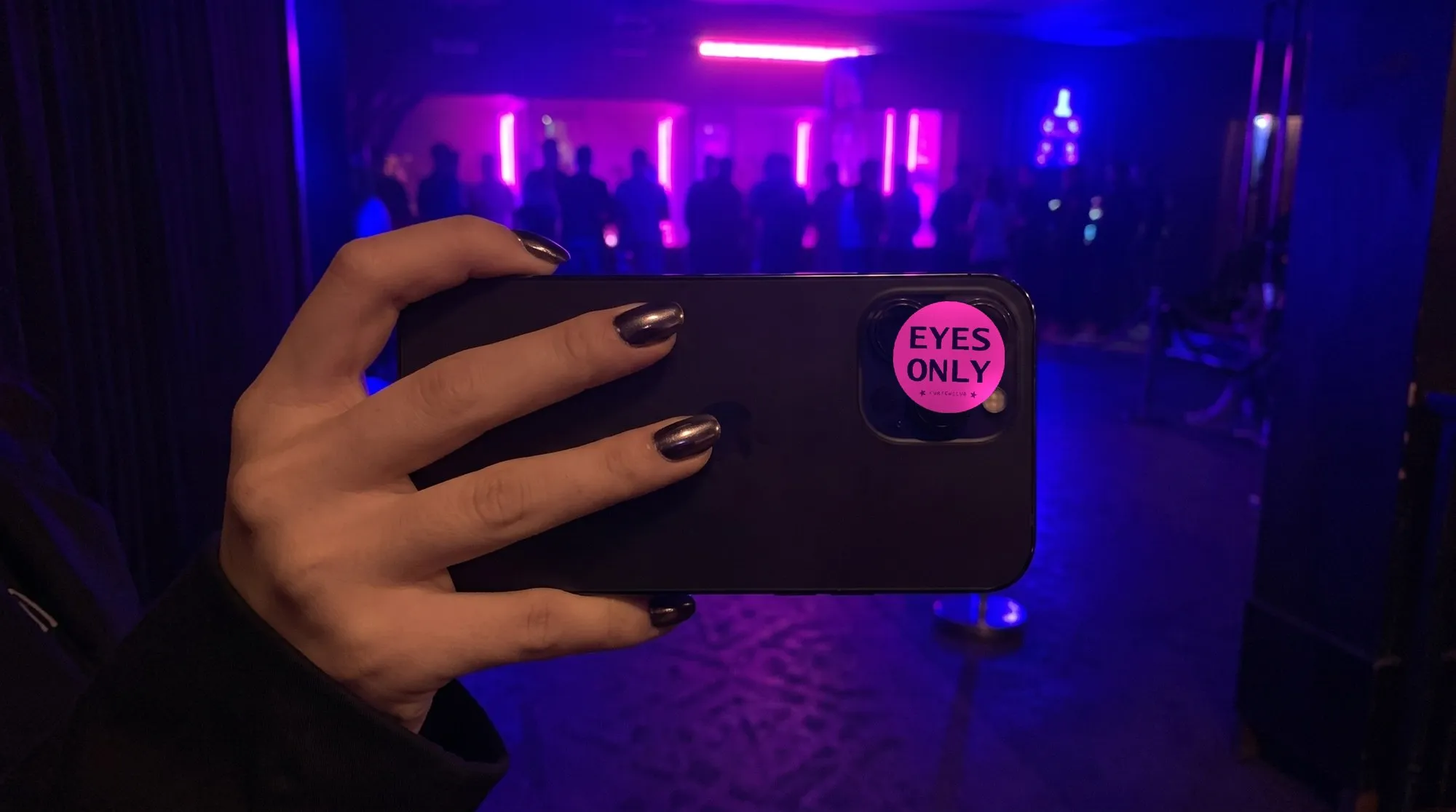 The back of a black phone held in a hand at the door of a dark club, with purple light behind. Stuck over the camera lenses is a round magenta sticker reading «EYES ONLY» in dark purple and, below it, the CURFEWCLUB mark between two stars.