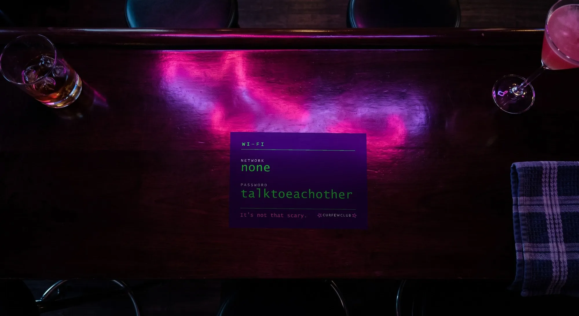 The dark counter of a bar seen from above, with magenta and purple reflections. In the middle, standing on it, a horizontal card reading «NETWORK: none» and «PASSWORD: talktoeachother» in green on a dark ground.