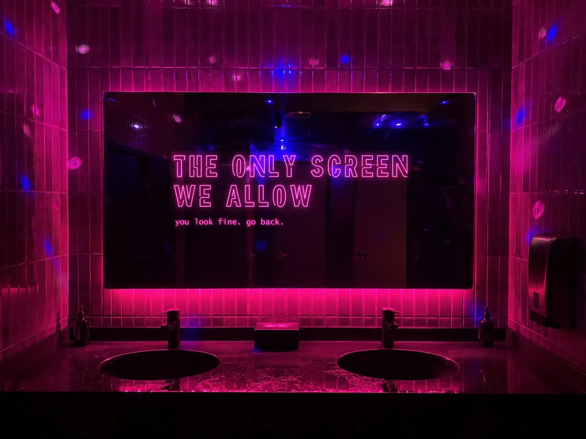 The mirror above the sinks in a club bathroom, with magenta tiles around it and a strip of pink LED underneath. On the glass, lit up, a pink neon sign on two lines — «THE ONLY SCREEN WE ALLOW» — and below it, smaller, «you look fine. go back.»