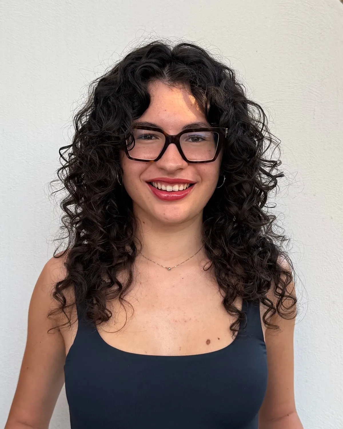 Sara sorride, in piedi davanti a un muro bianco: capelli ricci scuri e lunghi, occhiali dalla montatura squadrata e spessa, canotta blu scuro.